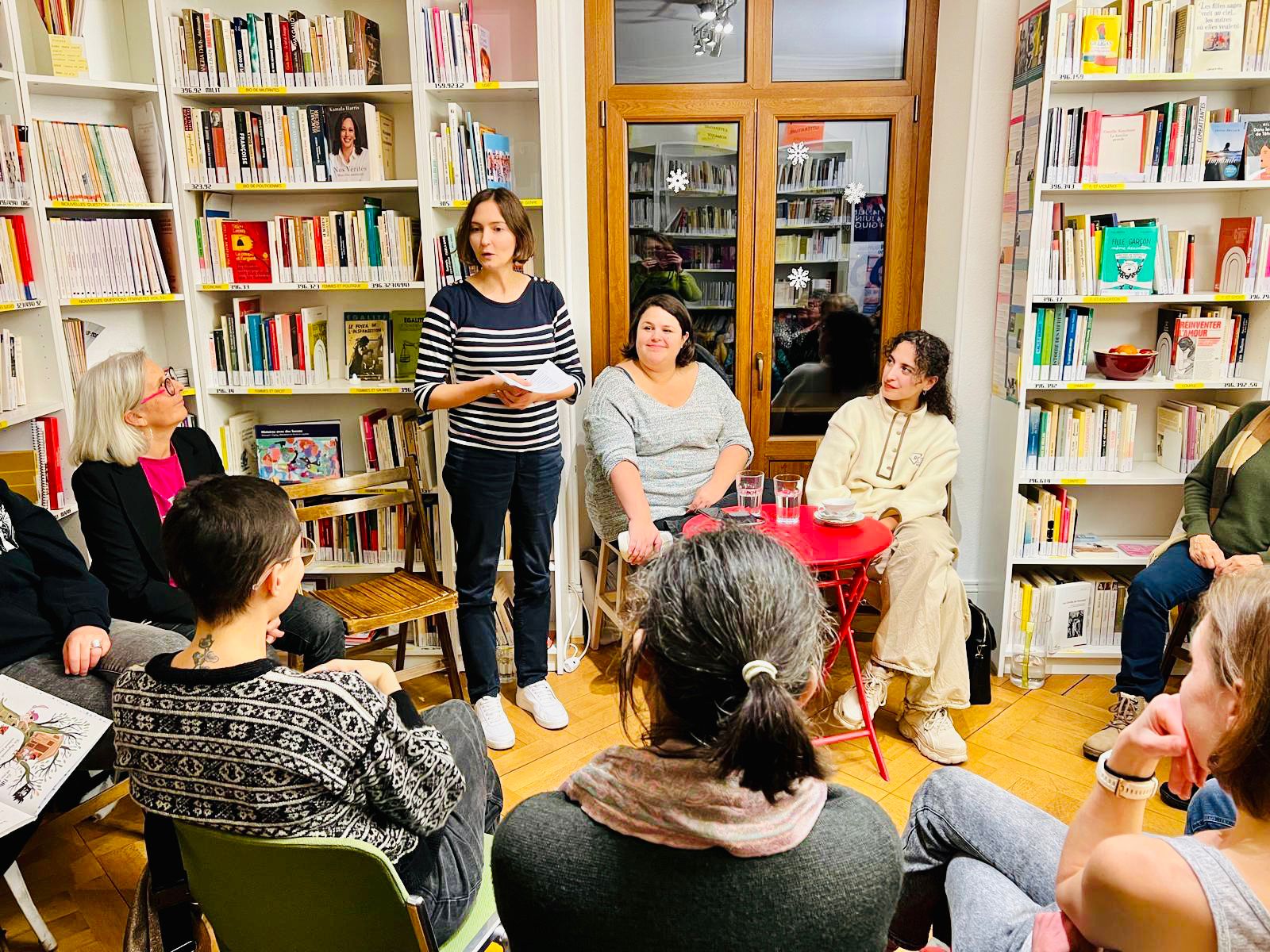 Lab_Evenement_RencontreEvasureKaziwa Rencontre avec Kaziwa Raim dans le cadre du club culturel l'Evasure en 2024. Sandra et Margaux ainsi que Kaziwa sont autour d'une table rouge. En find des bibliothèques remplies de livres. Kaziwa et Margaux regardent Sandra qui s'adresse au public. Le public est visible de dos au premier plan.